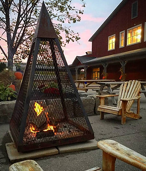 An enclosed fire pit surrounded by chairs with the market building in the background lighted up from inside 