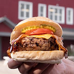 A person holding a juicy burger topped with bacon, cheese, tomatoes, and pickles from Country Cravings
